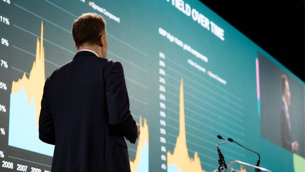 Speaker in suit on event stage with large LED screen showing financial data and charts