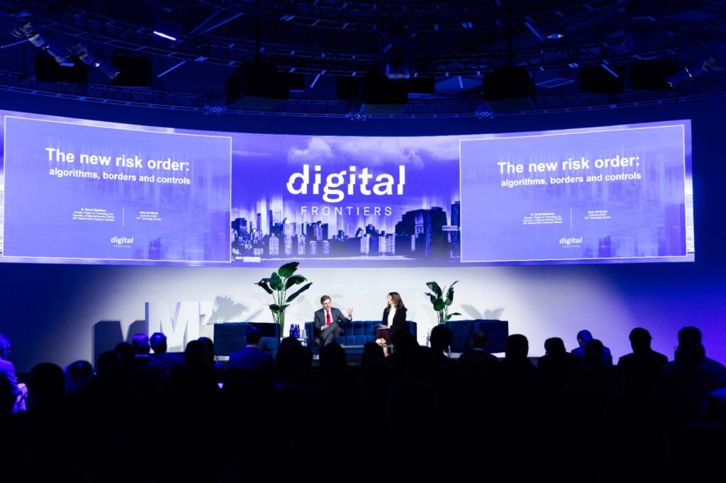 Conference stage with large curved screen and two speakers sat on a sofa on the stage