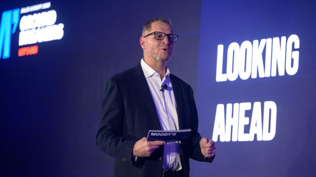Man in suit on stage holding speaker cards, in front of a screen that says Looking Ahead